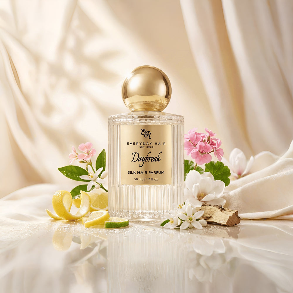 A bottle of Everyday Hair London Daybreak silk hair perfume is styled with white flowers, pink blossoms, lemon slices, and a piece of ginger on draped cream-colored fabric.
