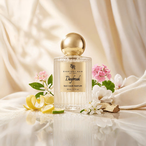A bottle of Everyday Hair London Daybreak silk hair perfume is styled with white flowers, pink blossoms, lemon slices, and a piece of ginger on draped cream-colored fabric.