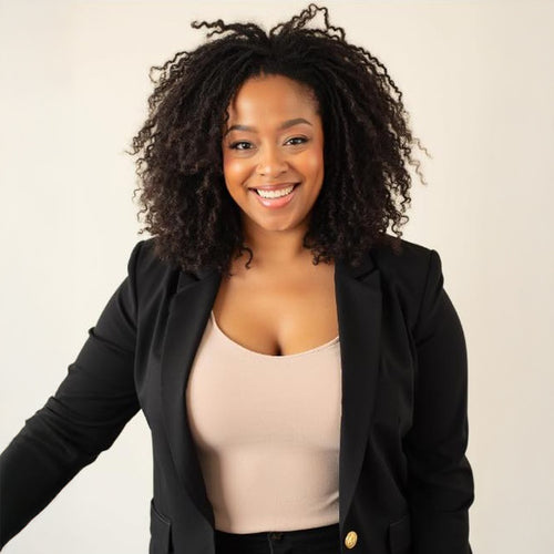 Smiling woman with natural curly hair wearing a black blazer over a beige top against a plain background.